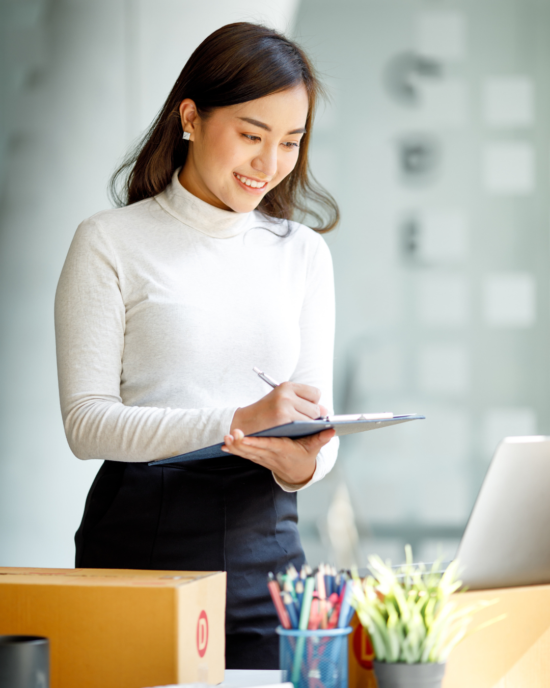 A smiling young Asian woman writing on a clipboard, surrounded by shipping boxes, symbolizing efficient order processing for logistics BPO services.