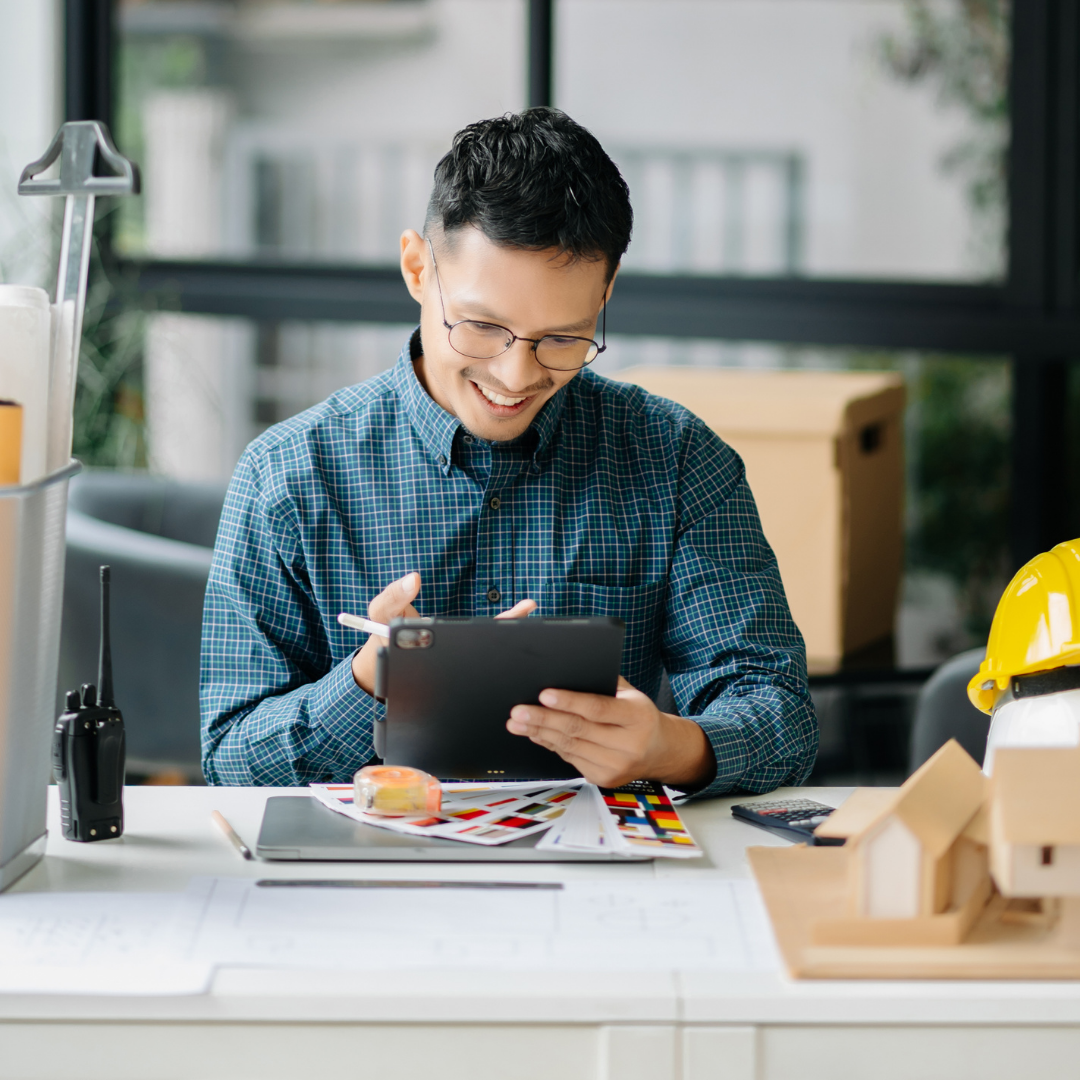 Strategic-Planning-for-Ecommerce-Management-Services Smiling Asian professional using a tablet at a desk with project plans, symbolizing detailed planning and strategic execution for ecommerce management services.